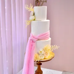 A dreamy and elegant three-tier engagement cake. Two tiers of blueberry and one of caramel are wrapped in pristine white fondant, accented with a flowing pink sash and delicate sugar flowers for a soft, romantic look.