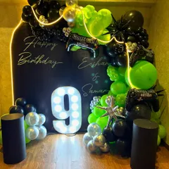 Get your game on with this epic gaming-themed ninth birthday party decor, featuring a bold black backdrop with neon-style light outlines and personalized "Happy Birthday" text. The setup is energized by a large light-up number '9' and a dynamic balloon arch in electric green, black, and silver, complete with gaming controller foil balloons.