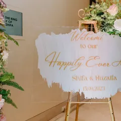 A personalized acrylic welcome sign for Shifa and Huzaifa's "Happily Ever After," elegantly scripted in gold and surrounded by lush floral arrangements.
