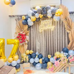 A stylish 17th birthday party decoration with a modern-boho theme. The setup includes a circular frame with a balloon garland, a 'Happy Birthday' neon sign, and large '17' foil balloons, accented with dried palm leaves.