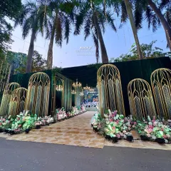 An opulent entrance for an outdoor wedding, featuring grand, gold cage-like structures, emerald green draping, and beds of pastel flowers.