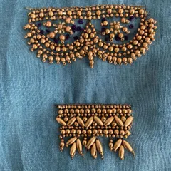 Two distinct embroidery patterns on blue fabric, showcasing different beading and threadwork techniques. The top design is a classic neckline pattern, while the bottom is a border style.