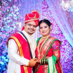 A classic portrait of the newlyweds on stage against a vibrant floral backdrop. The groom is wearing a traditional pheta (turban), and the bride is in a beautiful red and green saree, representing the complete Maharashtrian bridal look.