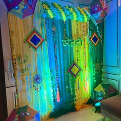 A Haldi or Mehndi backdrop with yellow and green drapes, marigold garlands, and ethnic umbrellas, illuminated with colorful lights.