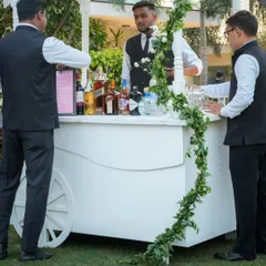 A charming white bar cart adorned with a simple garland of greenery and white roses, ready to serve celebratory drinks at the sundowner event.