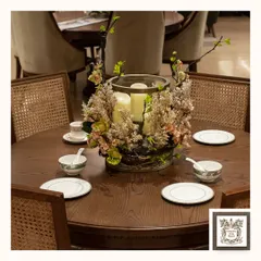 A close-up of a dining setting that feels both warm and elegant. The centerpiece, with its soft candlelight and artificial florals, sits on a dark wood table surrounded by classic wicker-back chairs, ready for an intimate gathering.