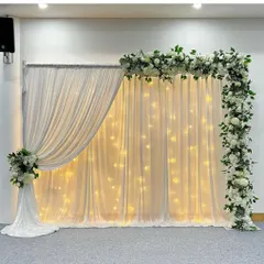 A classic and sophisticated backdrop made of white drapery and fairy lights. The look is completed with a beautiful white and green floral garland framing the top and side, perfect for an elegant celebration.