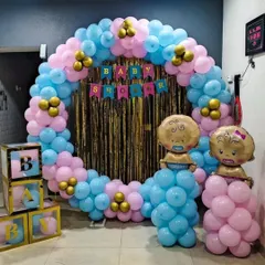 A classic baby shower decoration with a circular balloon arch in pink and blue, a gold shimmer curtain, and foil baby balloons. The transparent 'BABY' blocks are filled with matching balloons for a cohesive look.
