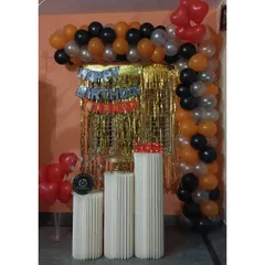 A simple and budget-friendly decor option for an at-home celebration. This setup uses an orange, black, and silver balloon garland with a gold tinsel curtain and pedestals for the cake.