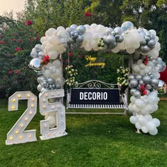 For a milestone 25th anniversary, I created this elegant outdoor swing decoration. The setup features a lush silver and white balloon arch, a custom 'Happy Anniversary' neon sign, and large marquee numbers, making it a beautiful focal point for photos in a garden or lawn setting.