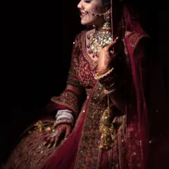 A dramatic and artistic portrait of a bride in her traditional red lehenga. The lighting highlights the rich details of her attire and her graceful pose.