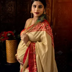 A graceful portrait of a mom-to-be in a cream and red silk saree, holding her bump with a peacock feather adding a touch of divine symbolism.