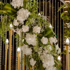 A detailed look at a floral arrangement on a modern arched backdrop. The combination of white hydrangeas, baby's breath, and green foliage against a curtain of crystal beads showcases our attention to detail.