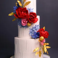 Where elegance meets indulgence. This three-tiered cake features a modern, textured white finish adorned with bold red and blue sugar flowers and striking golden leaves.