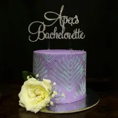 This purple bachelorette cake showcases a beautiful stenciling technique. The delicate silver leaf pattern on the side is complemented by a fresh white rose and a glittery custom cake topper.