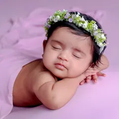 A peaceful baby in the 'chin on hands' pose, adorned with a beautiful floral crown against a soft lavender background.