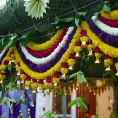 A close-up of a beautiful floral thoranam for a housewarming. This one is made with coconut leaves, colorful flowers, and small brass bells that jingle as guests arrive.