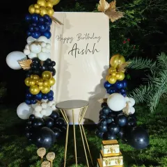 This outdoor setup features a modern, custom-printed backdrop panel with a personal birthday message. It's framed by an organic balloon garland in a stylish navy, white, and gold color scheme, perfect for garden or terrace parties.