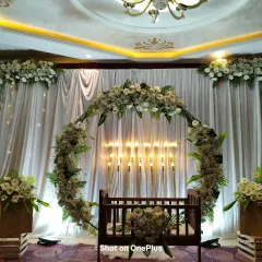 An elegant naming ceremony setup with a circular arch of white flowers and greenery. The traditional wooden cradle is placed in the center, with soft candle lighting in the background to create a warm and inviting atmosphere.