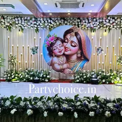 A grand and elegant stage for a baby shower with a Yashoda-Krishna theme. The backdrop is adorned with white flowers, and the stage is lined with a dense floral border.