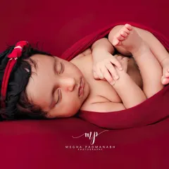 Another angle of the tucked-in pose, showcasing the baby's profile and the gentle curve of their back. The simple red backdrop makes the portrait pop.