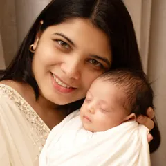 A glowing new mother smiles warmly at the camera while holding her peacefully sleeping newborn, a simple and beautiful portrait of the maternal bond.