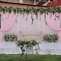 A soft and romantic stage setup perfect for an outdoor or daytime reception. The design uses flowing pink drapery, a clean white platform, and abundant greenery with white and pink florals for a fresh, whimsical feel.