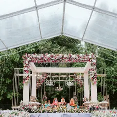 A breathtaking mandap setup inside a clear marquee tent, surrounded by lush greenery. The modern white structure was adorned with beautiful red and white florals and crystal chandeliers for a truly dreamy wedding ceremony.
