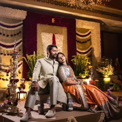 A quiet moment for the couple amidst their beautiful Muhurtham decor. The backdrop features a floral 'naamam' symbol, blending traditional elements with elegant, large-scale flower arrangements.
