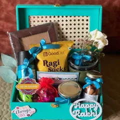 A vibrant turquoise Rakhi gift box with a cane-patterned lid. It's filled with snacks, a photo frame, and a 'Happy Rakhi' cutout.