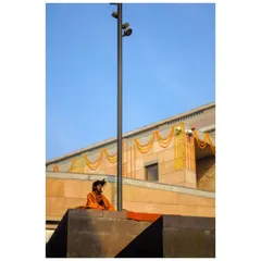 A sadhu in orange robes sits meditating against a modern building in Varanasi. The vibrant colors contrast with the stark geometry of the architecture.
