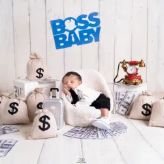 A series from a 'Boss Baby' themed newborn shoot. The setup includes a tiny armchair, a miniature briefcase, and prop money, creating a playful and humorous scene for a baby boy's first portraits.