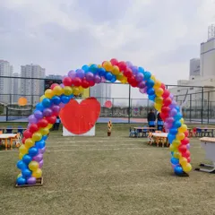 A large rainbow balloon arch set up outdoors in a park or school ground, perfect for a sports day or a large kids' party.