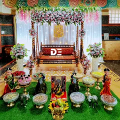 This Seemantham ceremony decor features a beautiful wooden swing (jhoola) decorated with flowers, set against a backdrop of white drapes and a traditional rangoli floor design.