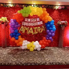 This is a custom birthday backdrop I created, featuring a circular rainbow balloon arch. It includes a 'Happy Birthday' banner and the child's name in foil balloons, set against a red floral wall.
