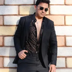 A classic portrait of a man in a sharp black suit, leaning against a brick wall. The lighting and pose create a confident and stylish look.