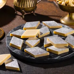Classic Kaju Katli for Grover Sweets. The macro focus and dark marble platter emphasize the smooth texture and silver leaf (vark) of this popular Indian sweet.