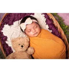 A close-up of a sleeping baby nestled in a basket with a teddy bear friend. The soft textures of the wrap and the bear create a cozy and comforting newborn portrait.