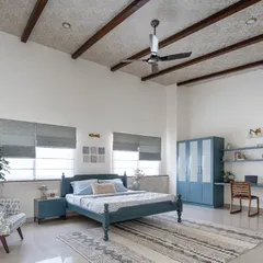 The master bedroom in the 'Spanish Country Home' features a serene blue color palette and a sloped roof with exposed rafters. Despite its spaciousness, the room feels cozy and intimate, complete with a custom study nook and wardrobe.
