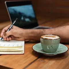 The perfect setup for a productive morning: a notebook, a pen, a laptop, and a fresh cup of our specialty coffee.