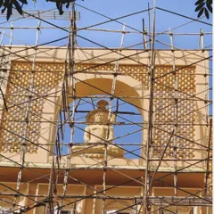 A close-up view of the Jain Upashray's facade during construction. This shot highlights the detailed craftsmanship of the stone statue and the surrounding jali work, framed by the scaffolding that supports its creation.