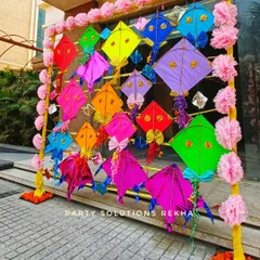 A creative photo backdrop using colorful kites for a spring or Makar Sankranti themed event. The pink paper pom poms add a soft, floral touch to the playful design.