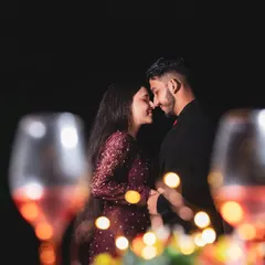 A creative shot of a couple's intimate moment, viewed through two wine glasses. The bokeh from the fairy lights in the background adds a magical, romantic feel to this night-time portrait.