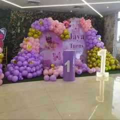 This is a butterfly-themed decoration for Jaya's first birthday. I used a purple and pink balloon arch, a custom backdrop, and a large light-up number '1' to create a vibrant and celebratory stage area.