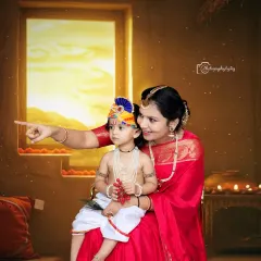 In this studio shot, a mother points towards a warm, glowing light from a window prop, sharing a story with her child dressed as Little Krishna. This setup creates a narrative feel, capturing a moment of discovery and wonder.