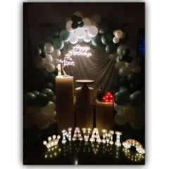 The lighting elements are key to this birthday setup for Navami. The marquee letters of her name are accented with a light-up crown and rainbow, creating a magical glow against the dark green and white balloons.
