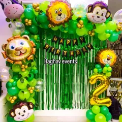 A fun jungle-themed birthday decoration. This setup includes a green foil curtain backdrop, a green balloon arch, and large foil balloons of jungle animals like lions and monkeys.