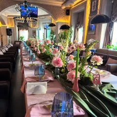 A beautifully styled long table for an intimate family gathering. The tablescape features a lush floral and moss runner, pink linens, and elegant blue glassware, creating a cozy and connected dining experience.