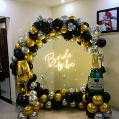 A glamorous bachelorette party backdrop featuring a balloon ring in a striking black, gold, and silver color scheme. The setup is accented with champagne and wine glass foil balloons for a fun, celebratory theme.