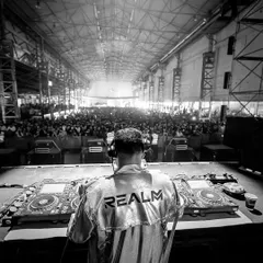 A shot from behind the decks at a massive warehouse-style festival. The raw, industrial setting combined with a huge crowd creates an intense and immersive techno experience.